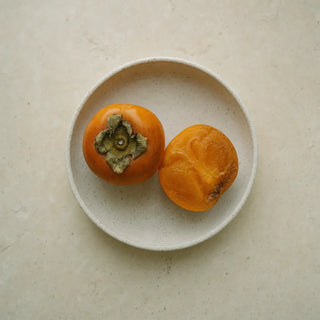 A whole ripe Japanese persimmon and a cut half in a cream ceramic dish