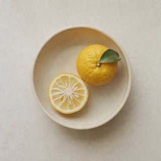 A whole yuzu with a leaf and a cut half in a cream ceramic dish